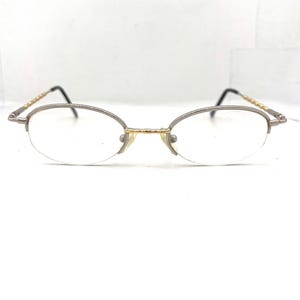 Op de afbeelding: Ovale bril met een zilverkleurig montuur en goudkleurige accenten. De glazen zijn helder en de pootjes zijn dun met een decoratief patroon. De bril heeft een klassiek, ingetogen ontwerp.