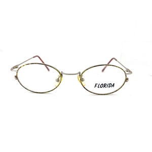 May include: Oval-shaped eyeglasses with a thin, silver-colored metal frame. The lenses are clear, and the frame has a tortoiseshell pattern around the lenses. The text "FLORIDA" is printed on the right lens. The temples are a dark red color.