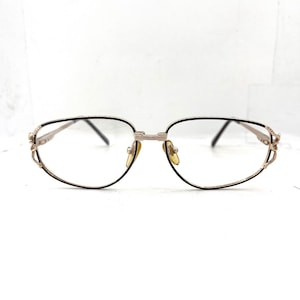 Könnte beinhalten: Goldfarbene Brille mit schwarzem Rand und klaren Gläsern. Die Fassungen haben dekorative Details an den Bügeln und am Steg. Die Brille ist vor einem weißen Hintergrund platziert.