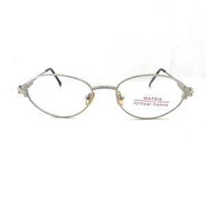 May include: Silver-toned oval eyeglasses with thin frames and delicate temple detailing. The lenses are clear, and the bridge features a small nose piece. The text "MATRIS optical frames" is visible on a sticker on the lens.
