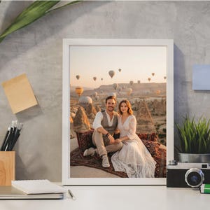 May include: A framed photograph featuring a couple seated on a patterned rug, with hot air balloons in the background. The photograph is in a white frame, and the scene is set against a backdrop of a scenic landscape.