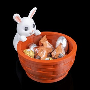 Op de afbeelding: Een oranje geweven mand gevuld met verpakte chocolaatjes en met folie bedekte eieren, met een wit konijnfiguur dat over de rand kijkt. Het konijn heeft roze binnenoren. Een paasdecoratie.