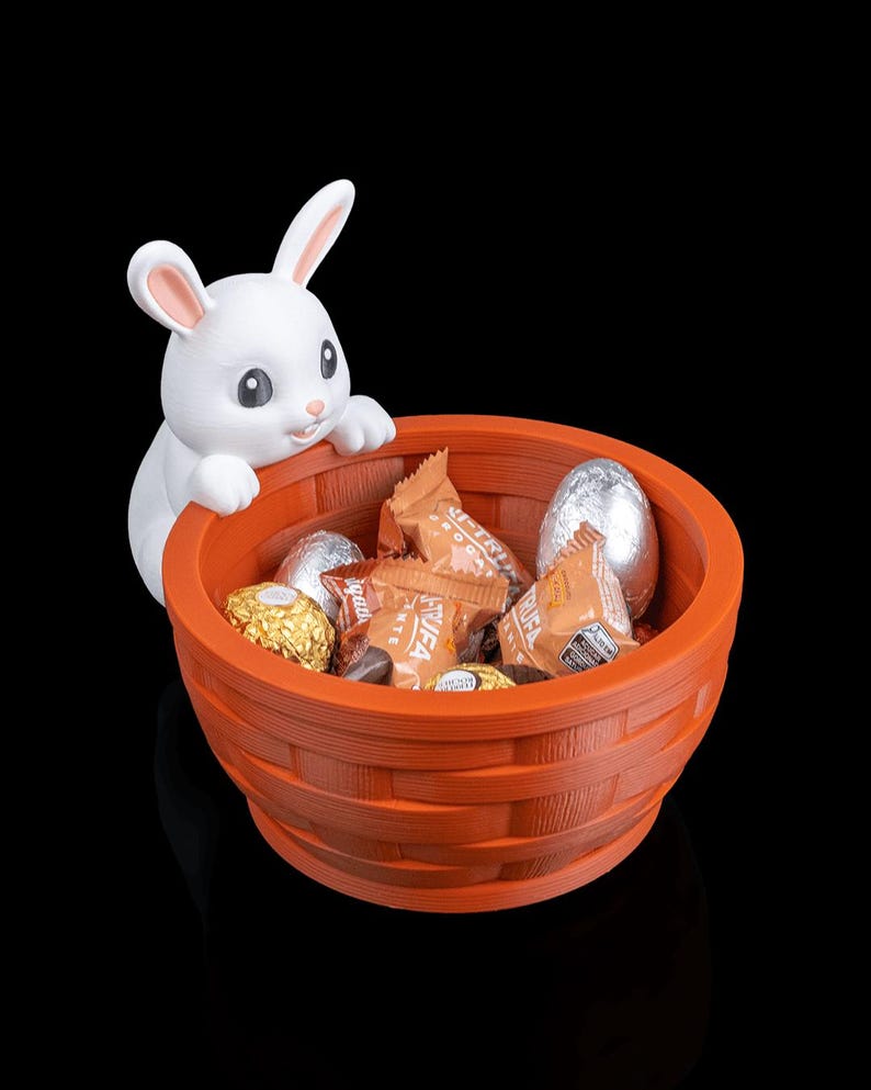Op de afbeelding: Een oranje geweven mand gevuld met verpakte chocolaatjes en met folie bedekte eieren, met een wit konijnfiguur dat over de rand kijkt. Het konijn heeft roze binnenoren. Een paasdecoratie.