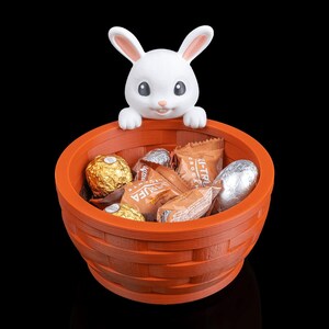 Op de afbeelding: Een oranje mand met een geweven ontwerp, gevuld met verpakte chocolaatjes, wordt bekroond door een wit konijnenfiguur. Het konijn heeft roze gevoerde oren en een lachend gezicht. De achtergrond is zwart.