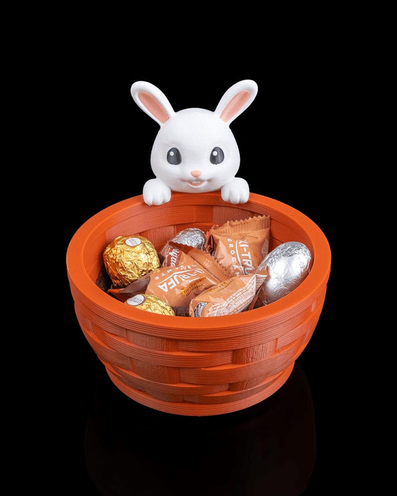 Op de afbeelding: Een oranje mand met een geweven ontwerp, gevuld met verpakte chocolaatjes, wordt bekroond door een wit konijnenfiguur. Het konijn heeft roze gevoerde oren en een lachend gezicht. De achtergrond is zwart.