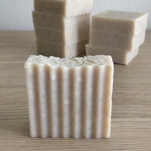 May include: Close-up of several rectangular bars of handmade soap. The front-most bar has vertical ridges. The soap is a light beige color and stacked in small piles. The background is a light-colored wall and a wooden surface.