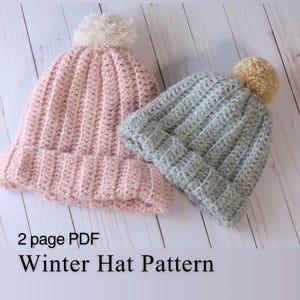 May include: Two handmade crochet winter hats. One is light pink with a cream-colored pom-pom, and the other is light gray with a tan pom-pom. The hats are ribbed and sit on a light wood surface. Text on the image reads "2 page PDF Winter Hat Pattern."