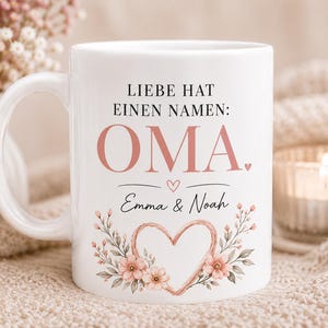 May include: White ceramic mug with a handle, featuring the text "LIEBE HAT EINEN NAMEN: OMA" in black and pink. Below, it reads "Emma & Noah" with a heart and floral design. The mug is decorated with a heart-shaped floral wreath.