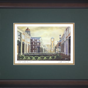 May include: Framed watercolor print of a university campus scene. The artwork depicts buildings with architectural details, a clock tower, and landscaping. The print is framed in a dark brown wood frame with a green mat, and the title "UMKC Golden Dome" is visible.