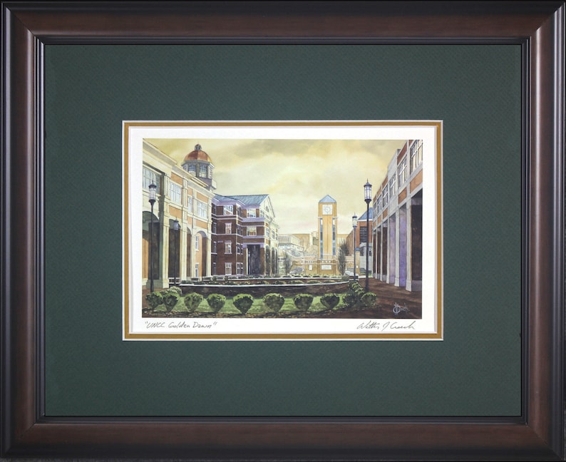 May include: Framed watercolor print of a university campus scene. The artwork depicts buildings with architectural details, a clock tower, and landscaping. The print is framed in a dark brown wood frame with a green mat, and the title "UMKC Golden Dome" is visible.