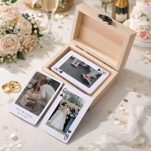 May include: A light wooden box with a hinged lid contains custom playing cards with wedding photos. Two cards are displayed, one with the names "Zane & Caliese" and the date "April 19, 2023." Gold wedding rings and a bouquet of white roses are in the background.