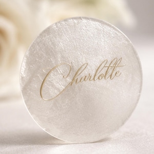 May include: A circular place card, translucent with a pearlescent sheen, featuring the name "Charlotte" in gold script. The card's design suggests a refined aesthetic, suitable for weddings or formal gatherings. The diameter is approximately 7 cm.