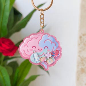 Könnte beinhalten: Ein rosa-blaues Gehirn-Schlüsselanhänger mit einer goldfarbenen Kette. Das Design zeigt medizinische Symbole. Ein lustiges Accessoire für Medizinstudenten oder Angehörige der Gesundheitsberufe.