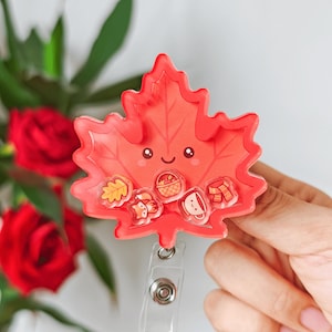 Puede incluir: Un portatarjetas de acrílico con forma de hoja de arce roja que presenta una cara sonriente y cinco pequeños dijes flotantes en su interior, incluyendo una hoja amarilla, un zorro, una cesta de manzanas, una taza blanca y una bufanda.