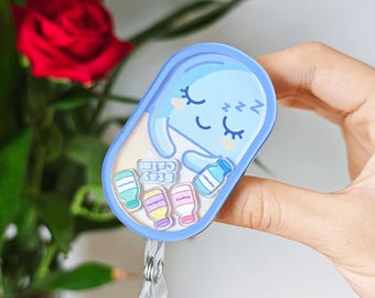May include: A light blue oval badge reel featuring a cartoon pill character with closed eyes and sleeping symbols. Inside the clear casing are small floating charms shaped like medicine bottles and the text Stay Calm in white lettering.