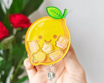 Puede incluir: Un portatarjetas con forma de limón amarillo con una hoja verde y una cara sonriente. El portatarjetas tiene un clip de plástico transparente y presenta pequeñas imágenes de rodajas de limón, postres y bebidas.