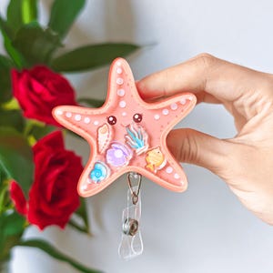 May include: A peach-coloured starfish badge reel featuring a clear window filled with small, floating ocean-themed charms including a seashell, a purple clam with a pearl, blue seaweed, and a yellow fish, finished with a metal clip and clear strap.