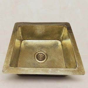 Op de afbeelding: Een vierkante wastafel van gehamerd messing met een metallic gouden afwerking. De bak heeft een getextureerd, gehamerd oppervlak over de gehele binnenzijde en de brede rand, met een ronde afvoer centraal in de bodem van de wastafel.