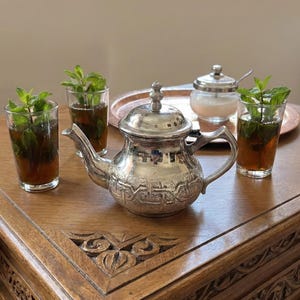 Könnte beinhalten: Ein silbernes Teeservice mit dekorativem Muster, einschließlich einer Teekanne, einer Zuckerdose und drei Gläsern Tee mit Minzzweigen. Das Set ist auf einem Holztisch mit geschnitzten Details und einem Kupfertablett angeordnet.