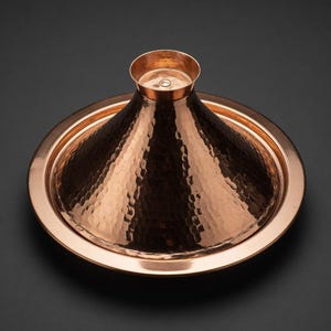Peut inclure: Un tajine en cuivre martelé doté d'un couvercle conique et d'une large base circulaire. La surface métallique présente une texture alvéolée distincte sur tout l'extérieur, reflétant la lumière sur la forme incurvée, surmontée d'une petite poignée.