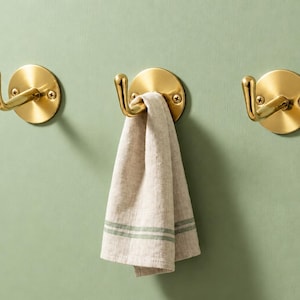 Peut inclure: Trois patères murales circulaires en métal doré avec une finition polie, chacune dotée d'un bras incurvé et de deux vis de fixation apparentes. Une serviette en mélange de lin beige avec deux fines rayures vertes est suspendue au crochet central.