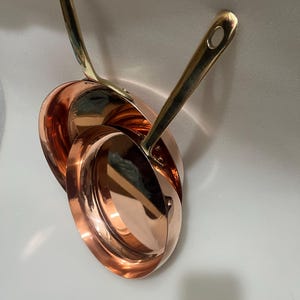 May include: Two polished copper frying pans with long, slender brass handles. The pans feature a reflective, metallic finish and are nested together, showcasing their circular shape and smooth, curved sides with a bright, warm orange-brown hue.