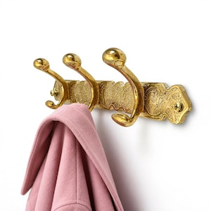Peut inclure: Un porte-manteau mural en métal doré doté de trois crochets incurvés et d'une plaque arrière rectangulaire ornée de motifs en relief complexes. Un manteau rose clair uni est suspendu au crochet situé à l'extrémité gauche.