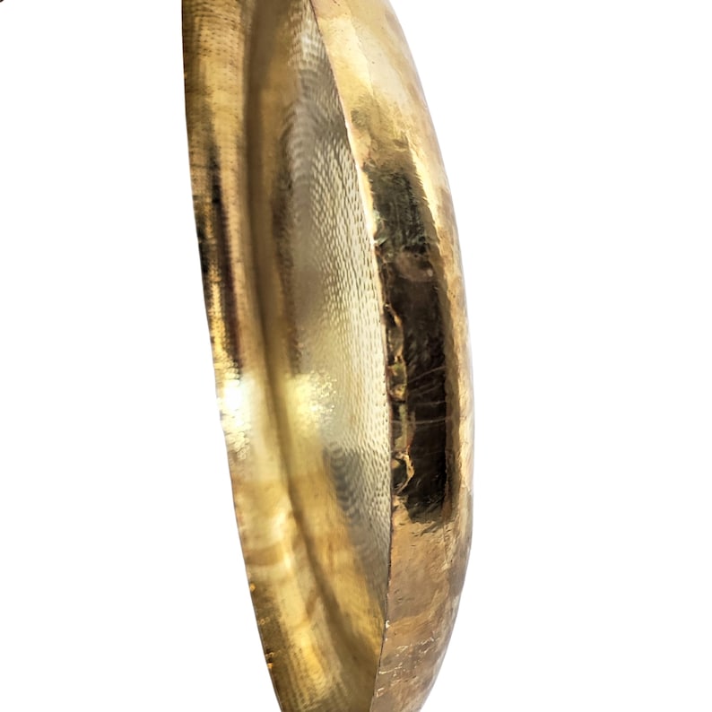 May include: Close-up of a gold-colored, hammered metal bowl. The bowl has a rounded shape with a textured surface and a shiny finish. The rim of the bowl is visible, showing the thickness of the metal. The background is white.