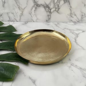 May include: A gold-colored, round decorative tray with a textured surface. The tray has a slightly raised edge and a hammered metal appearance. The background is a white marble pattern with green leaves.