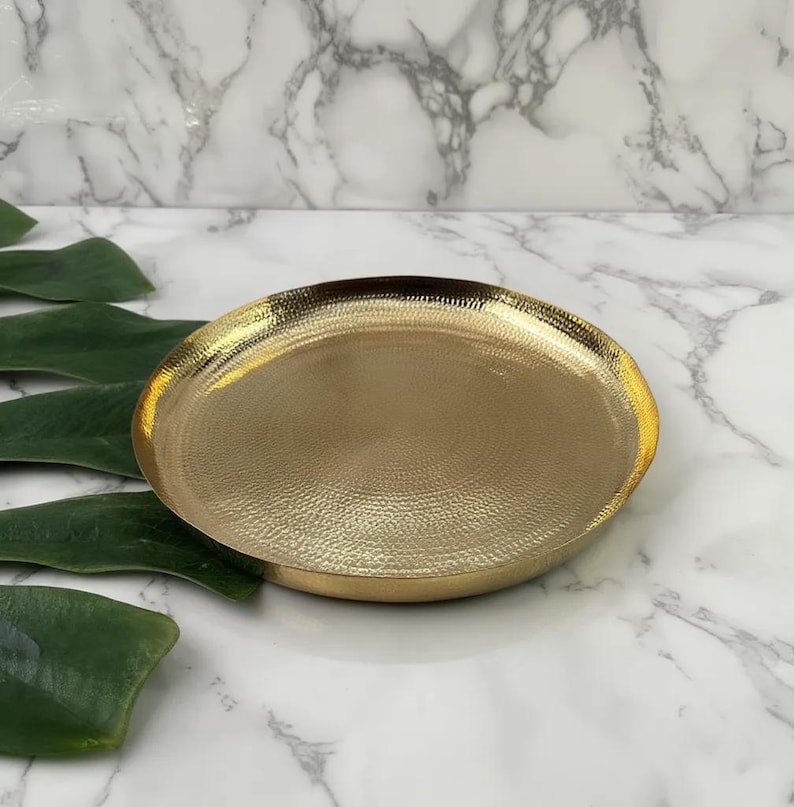May include: A gold-colored, round decorative tray with a textured surface. The tray has a slightly raised edge and a hammered metal appearance. The background is a white marble pattern with green leaves.