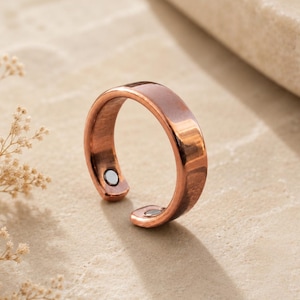 Op de afbeelding: Een gepolijste koperkleurige ring met een open bandontwerp. De binnenkant van de ring is voorzien van twee kleine, ronde, zilverkleurige magnetische inzetstukken aan beide uiteinden van de band, wat een contrast vormt met de koperen afwerking.