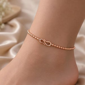 Op de afbeelding: Een enkelbandje van roségoud met een fijne ketting van kleine, uniforme, bolvormige kraaltjes. Het enkelbandje wordt vastgemaakt met een opvallende S-vormige haaksluiting, die als centraal middelpunt van het sieraad fungeert.