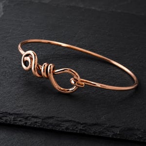 Può includere: Un bracciale rigido in metallo color oro rosa con finitura lucida. Il bracciale è realizzato con un filo continuo che forma un dettaglio decorativo a nodo vorticoso sulla parte anteriore, creando una forma circolare coesa e singola.