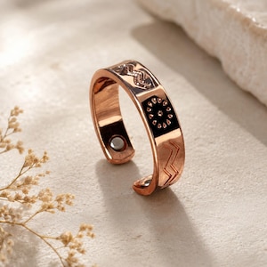 Puede incluir: Un anillo abierto de tono cobre con un acabado metálico pulido. La superficie exterior está decorada con patrones grabados que incluyen líneas en zigzag y un motivo de sol circular. Un pequeño imán circular es visible en la banda interior.