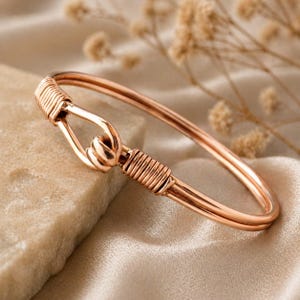 Puede incluir: Una pulsera brazalete de oro rosa con cierre de gancho y ojo. La pulsera presenta un diseño envuelto en alambre en el cierre y en el lado opuesto. La joya se exhibe sobre una superficie de tonos neutros con acentos florales secos.