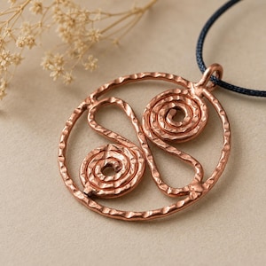 Könnte beinhalten: Ein kreisförmiger Kupferanhänger mit gehämmertem Muster und zwei innenliegenden Spiraldesigns. Der Anhänger hängt an einer dunkelblauen Kordel und weist eine metallische Oberfläche mit einem warmen rotbraunen Farbton auf.