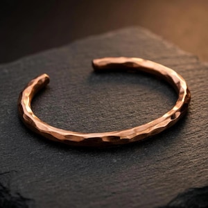 Könnte beinhalten: Ein Kupfer-Armreif mit einer gehämmerten, strukturierten Oberfläche. Der Armreif hat ein offenes Design, das eine kreisförmige Form mit einer Lücke an der Vorderseite bildet. Das Metall hat einen warmen, rötlich-braunen Farbton mit Reflexionen.