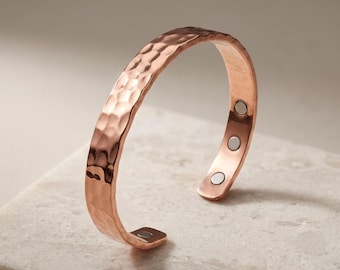 Puede incluir: Una pulsera tipo brazalete de metal en tono cobre con una superficie exterior martillada y texturizada. El interior de la banda abierta muestra tres pequeños insertos magnéticos circulares de color plateado cerca de los extremos.