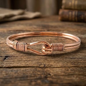 Op de afbeelding: Een koperkleurige armband met een uniek knoopontwerp. De armband heeft gewikkeld draaddetail aan weerszijden van de knoop, wat textuur toevoegt. De armband wordt gepresenteerd op een houten oppervlak.