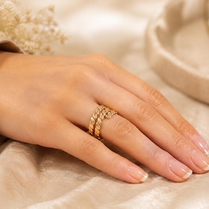 May include: A gold ring featuring a double-band design with a twisted rope texture. The ring is worn on the ring finger, showcasing a polished metallic finish and a continuous, interconnected pattern that wraps around the finger in two distinct rows.