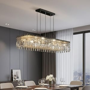 Op de afbeelding: Een rechthoekige kristallen kroonluchter hangt boven een eettafel. De kroonluchter heeft een zwart frame en is versierd met talrijke kristallen. De eetkamer heeft een donkere houten tafel, grijze stoelen en een neutraal kleurenschema.