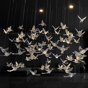 Puede incluir: Una lámpara de araña moderna con numerosos adornos de vidrio transparente en forma de pájaros suspendidos del techo. Los pájaros están iluminados, creando un efecto brillante sobre un fondo oscuro. El diseño incluye un sofá y una mesa pequeña.