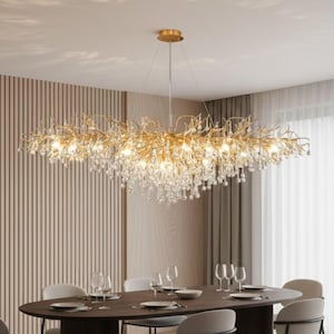 Op de afbeelding: Een goudkleurige kroonluchter met een takachtig ontwerp, versierd met talrijke heldere kristallen druppels. Hij hangt boven een donkerbruine ovale eettafel, gedekt met borden, glazen en bestek. De kamer heeft een neutraal kleurenpalet.