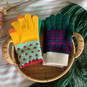 May include: Two pairs of colourful gloves in a woven basket. One pair is yellow with orange fingertips and a blue floral pattern. The other pair is green with a red and blue plaid pattern and white cuffs. The basket sits on a striped surface.