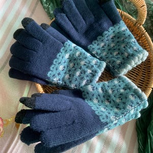 May include: A pair of navy blue knitted gloves with a light blue and white patterned cuff. The fingertips are black, suitable for touchscreens. The gloves are on a wicker basket and a striped pink and white fabric.