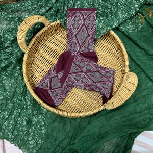 Op de afbeelding: Een paar sokken met patroon in een rieten mand. De sokken hebben een herhalend ruitontwerp in bordeauxrood en groen. De mand heeft aan elke kant een handvat en staat op een groene stoffen achtergrond.