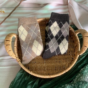 May include: Two pairs of argyle patterned socks in a woven basket. One pair is tan, brown, and cream, and the other is dark grey, light grey, and cream. The basket is made of woven wicker and has two handles.