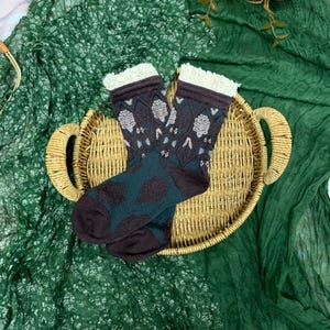 May include: Patterned socks in dark teal and brown, with white lace trim, resting on a wicker tray. The socks are displayed on a green fabric background, with flowers. The socks are designed with a geometric pattern.
