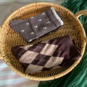 Könnte beinhalten: Zwei Paar Socken in einem Weidenkorb. Ein Paar ist braun mit weißen Blumenmustern, das andere ist braun mit einem beigen und braunen Rautenmuster. Der Korb liegt auf einer gestreiften Oberfläche.