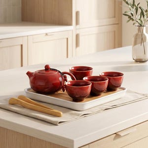 May include: A red ceramic tea set, comprising a teapot and four cups, displayed on a white tray with a bamboo insert. Includes bamboo tongs and a black decorative string. A traditional tea set.